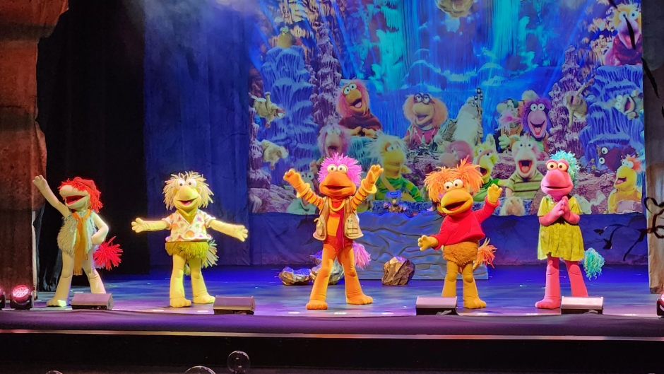  Five colorful puppet characters perform on stage under bright lights, with a detailed, vibrant backdrop behind them.